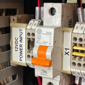 Close-up of an electrical panel with a labeled circuit breaker, orange switch, and wires. 430mm x 20mm Adhesive PVC Strips are used for clear labeling near the 12VDC POWER INPUT ON/OFF switch and terminal blocks.